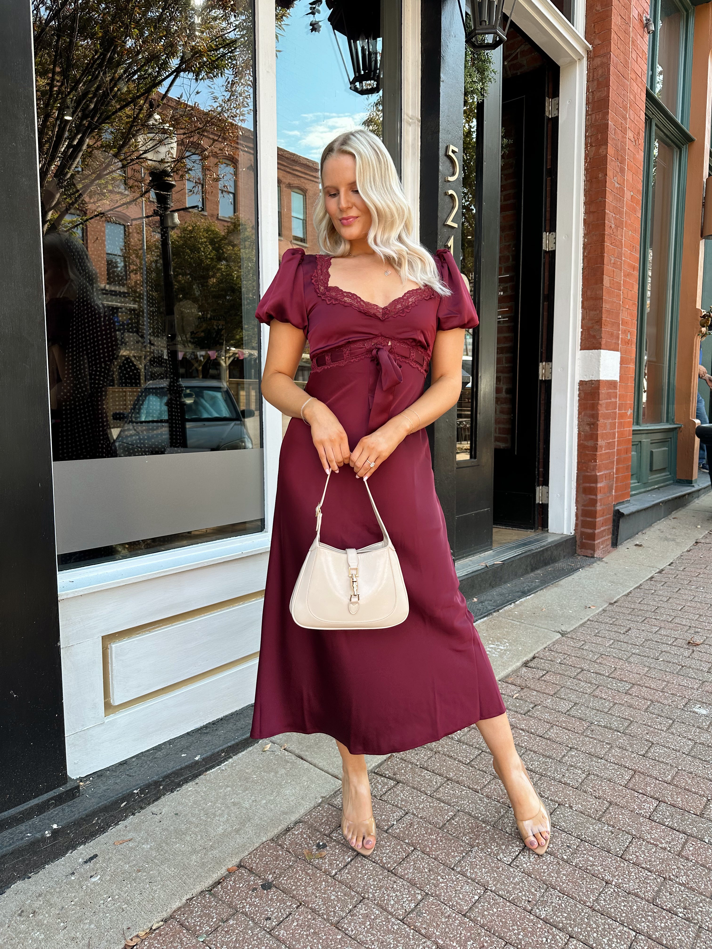 Woman wearing burgundy satin midi dress with puff sleeve, and lace detailing.