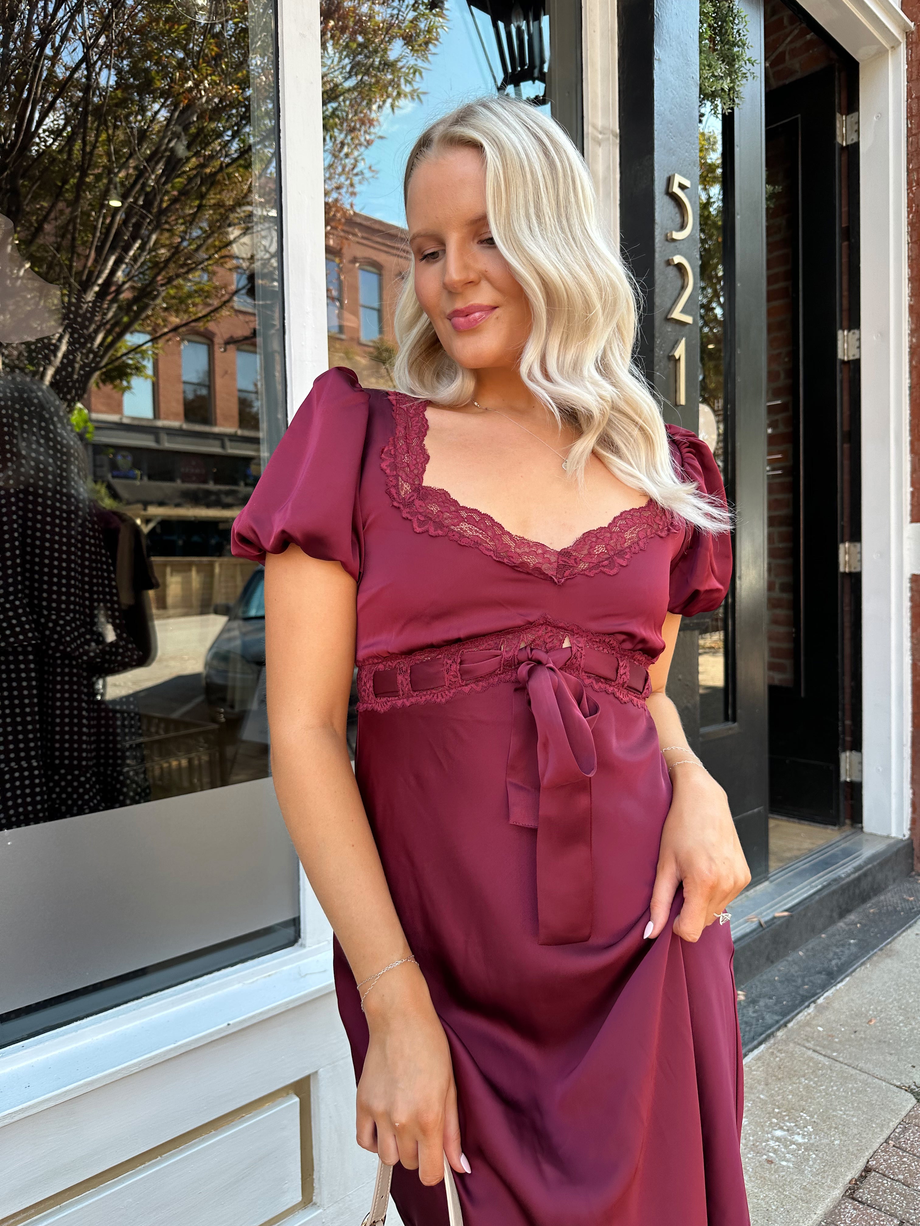 Woman wearing burgundy satin midi dress with puff sleeve, and lace detailing.
