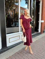 Woman wearing burgundy satin midi dress with puff sleeve, and lace detailing.