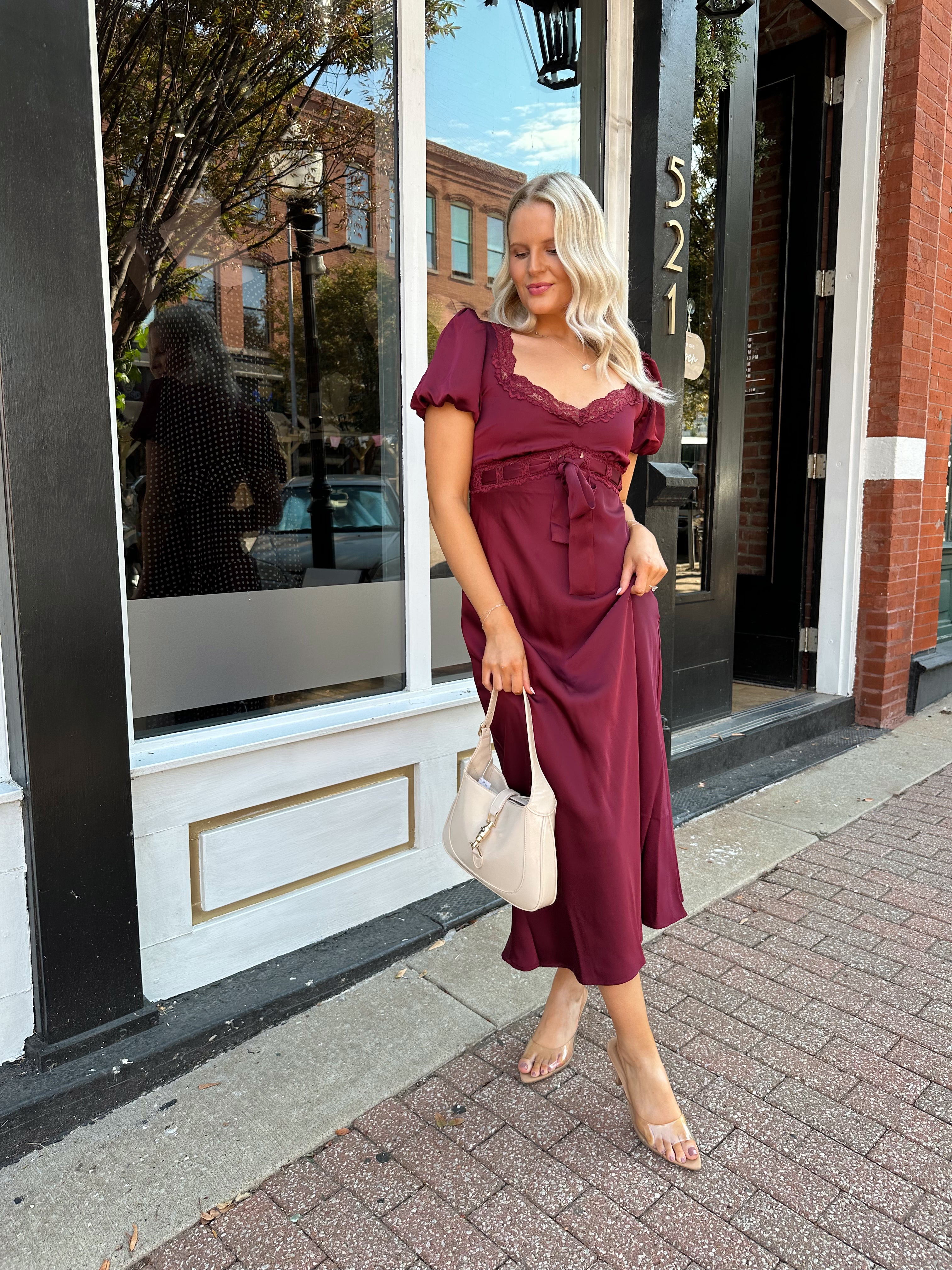 Woman wearing burgundy satin midi dress with puff sleeve, and lace detailing.
