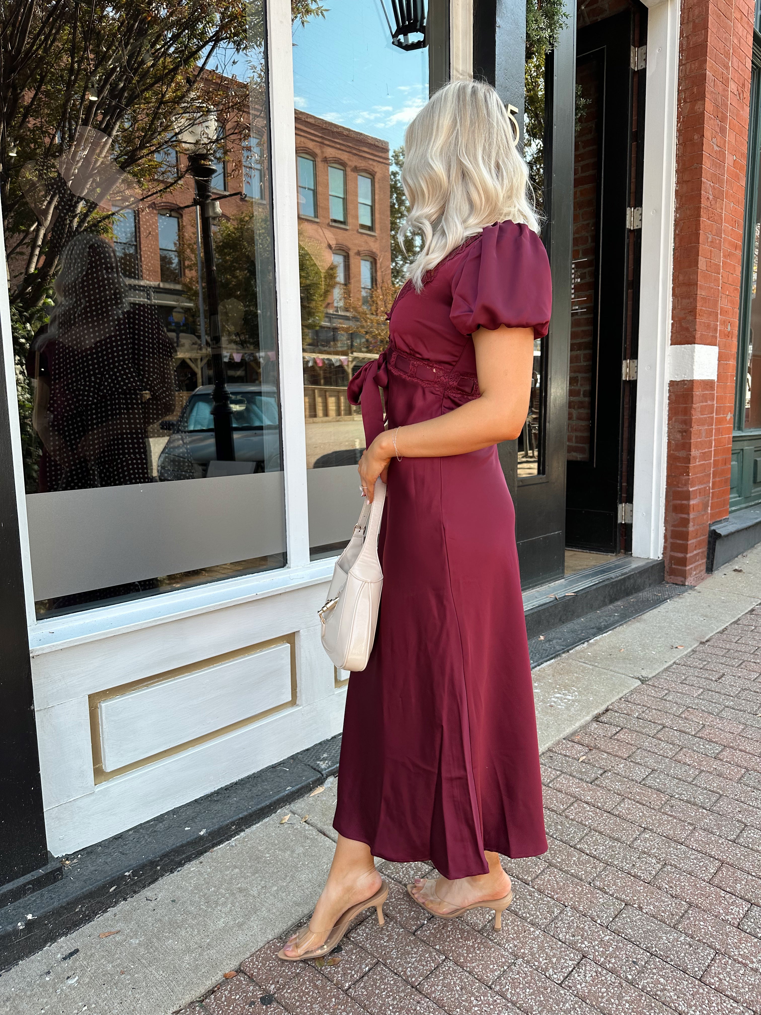 Woman wearing burgundy satin midi dress with puff sleeve, and lace detailing.