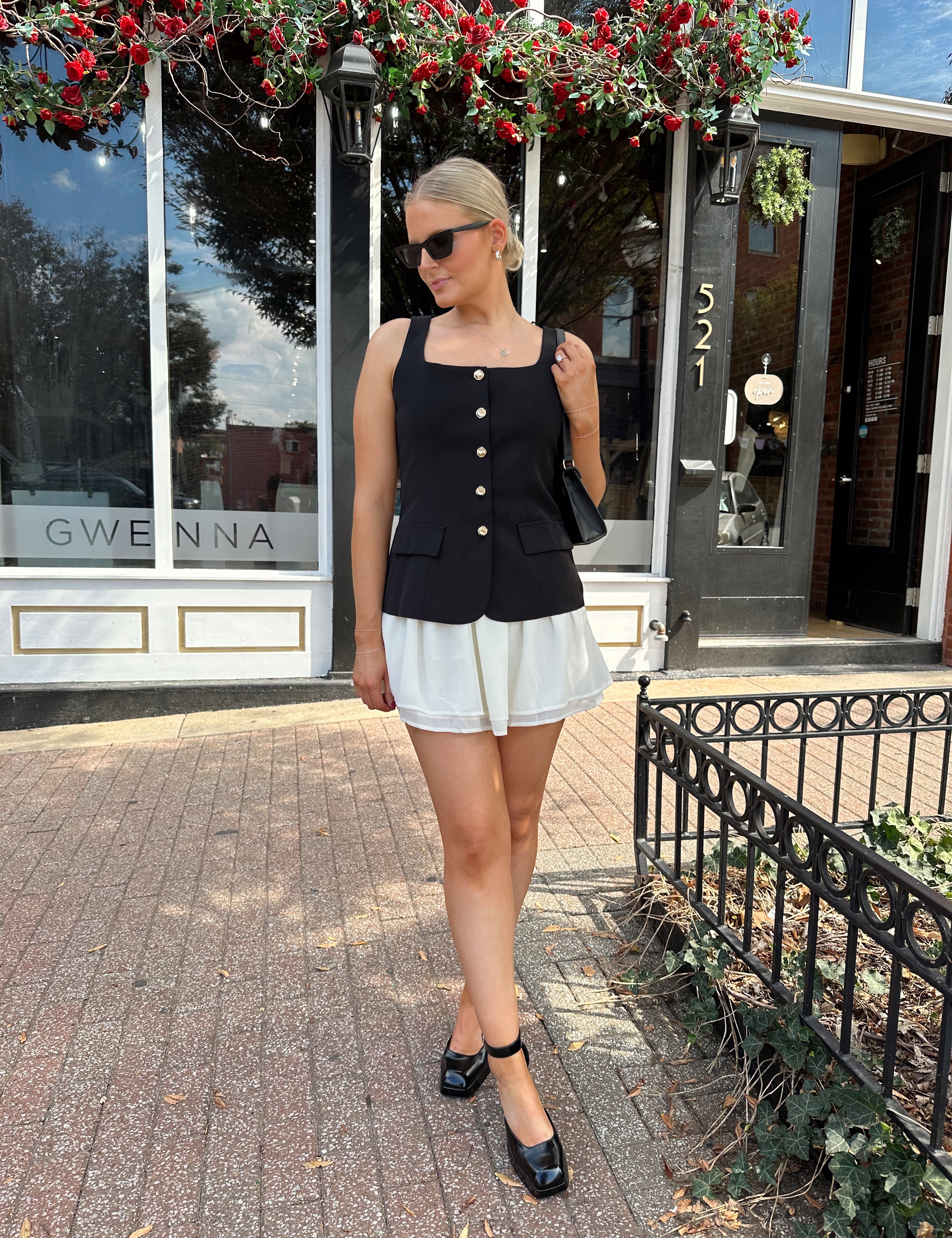 Woman wearing black tailored mini dress with white overlay poking out and gold button closure. 