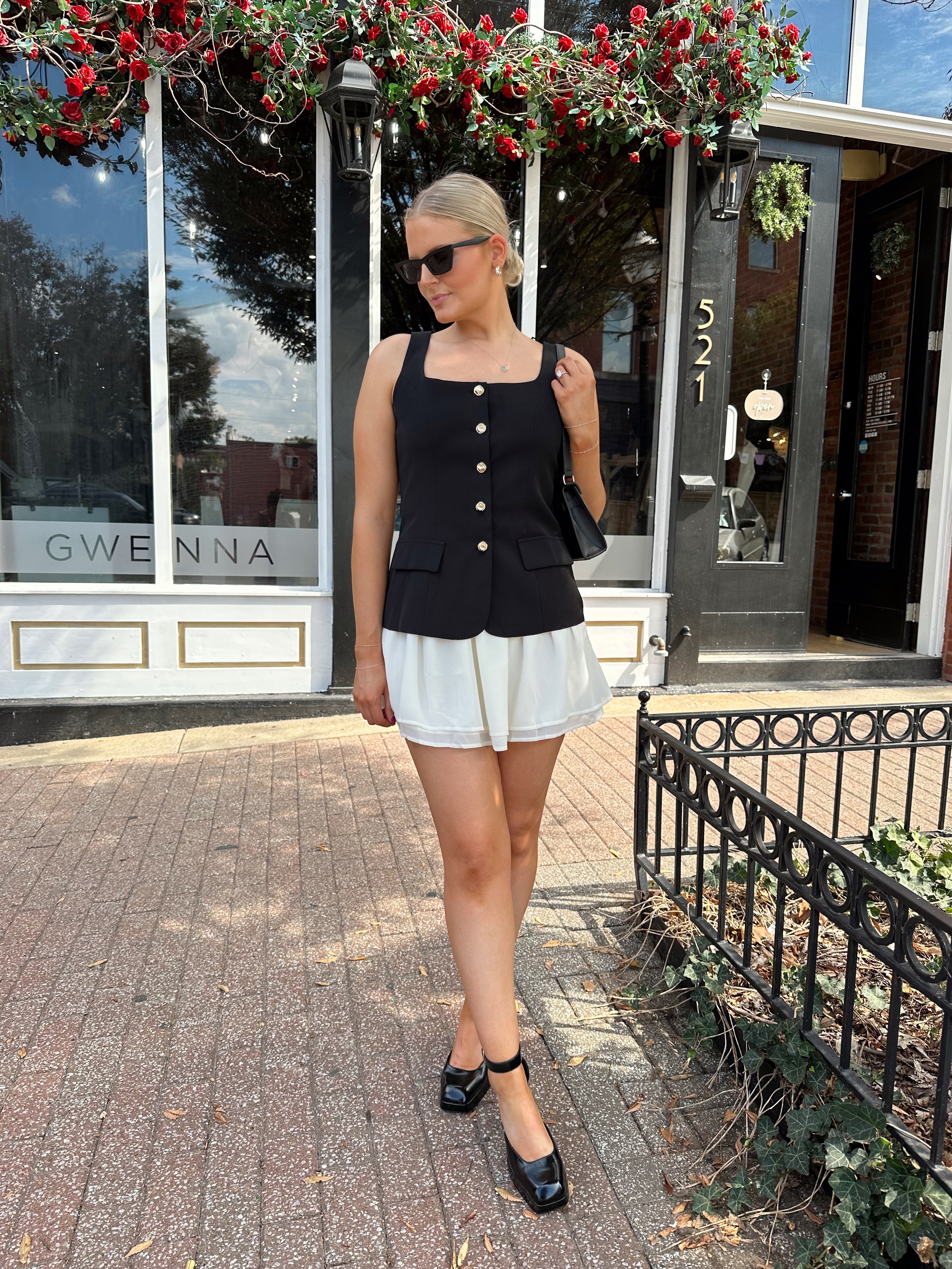 Woman wearing black tailored mini dress with white overlay poking out and gold button closure. 
