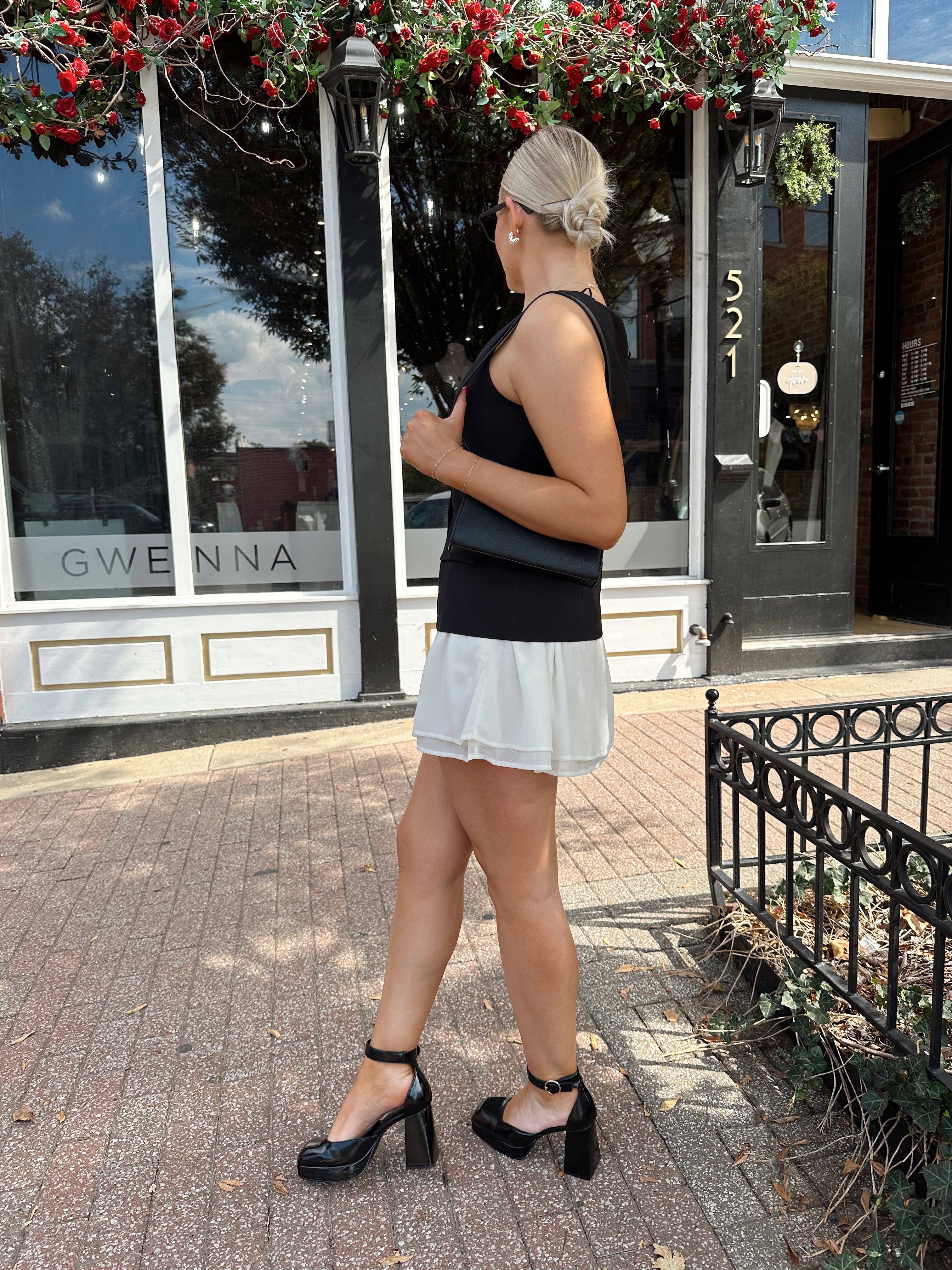 Woman wearing black tailored mini dress with white overlay poking out and gold button closure. 