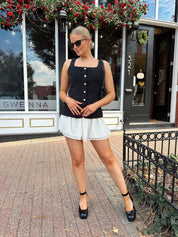 Woman wearing black tailored mini dress with white overlay poking out and gold button closure. 