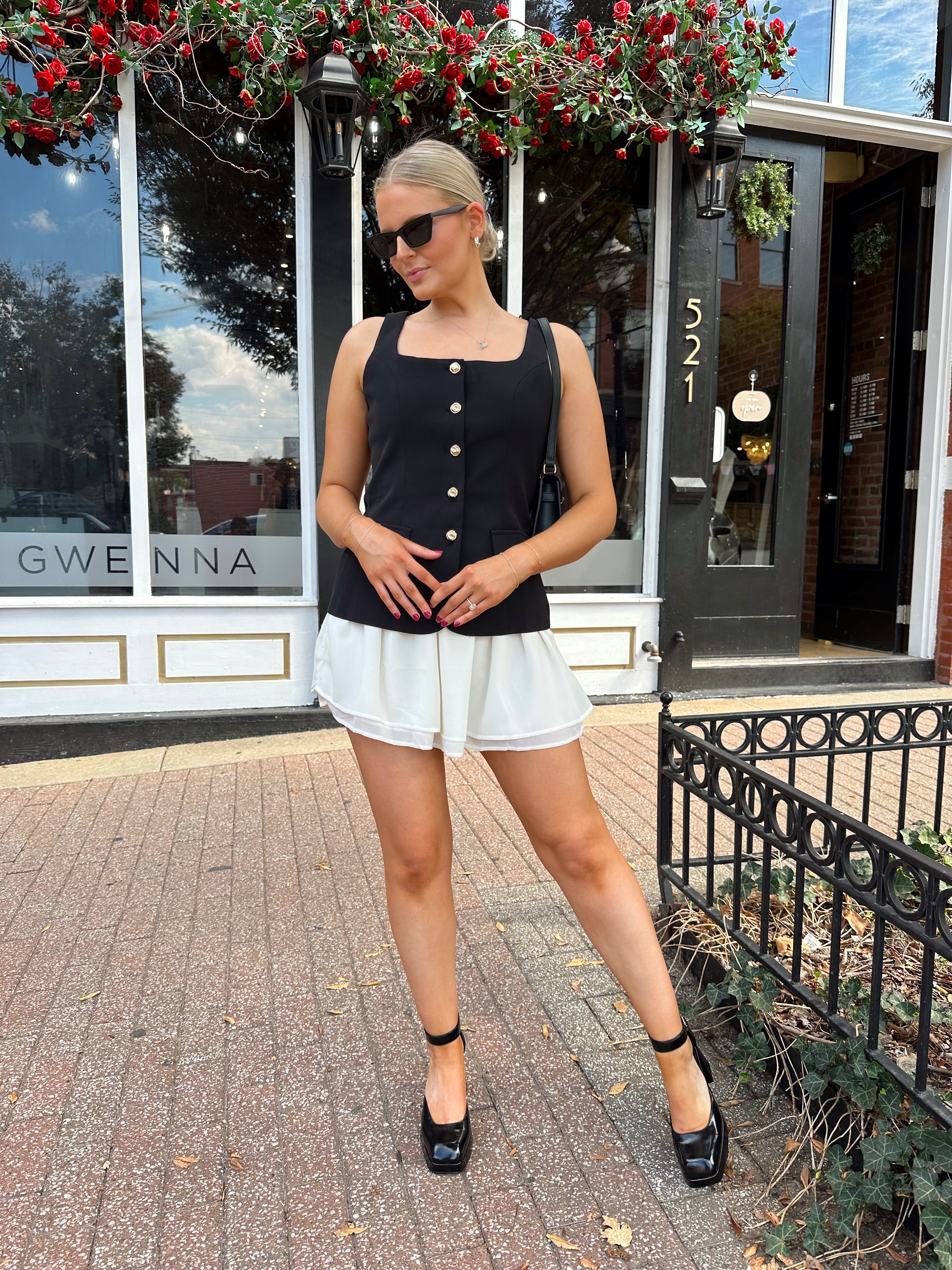 Woman wearing black tailored mini dress with white overlay poking out and gold button closure. 