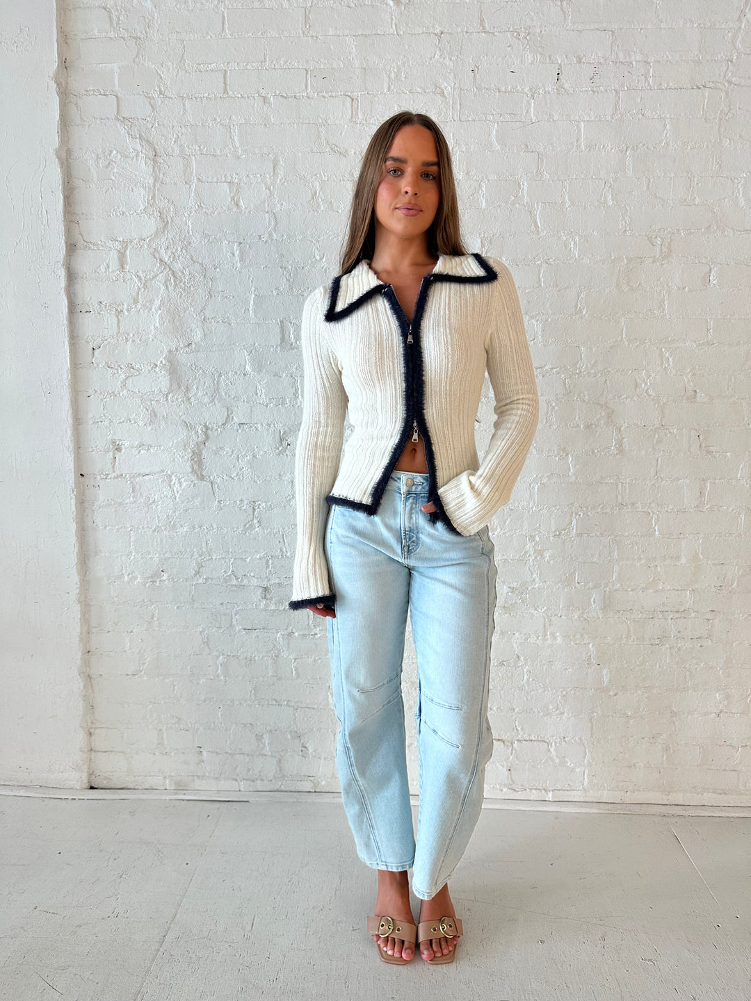 Woman wearing ivory knit long sleeve top with double zipper closure, and light wash barrel jeans.