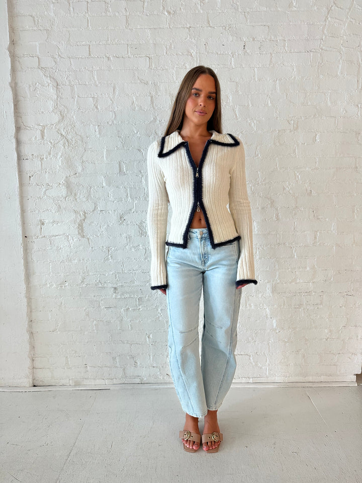Woman wearing ivory knit long sleeve top with double zipper closure, and light wash barrel jeans.