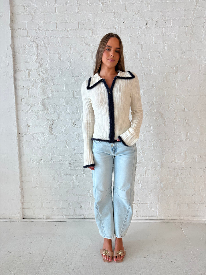 Woman wearing ivory knit long sleeve top with double zipper closure, and light wash barrel jeans.