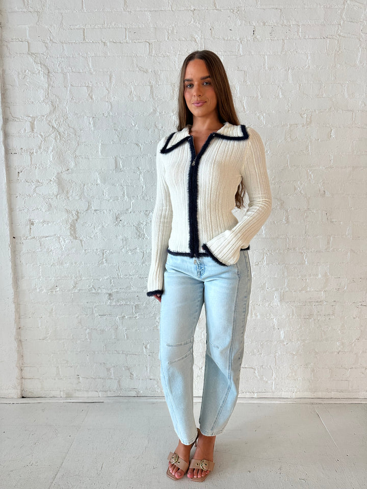 Woman wearing ivory knit long sleeve top with double zipper closure, and light wash barrel jeans.