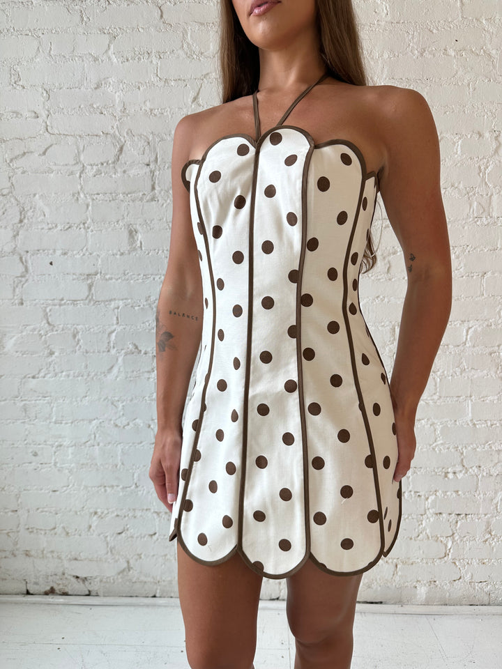 Woman wearing cream and brown polka dot mini dress with scallop neckline and hem.
