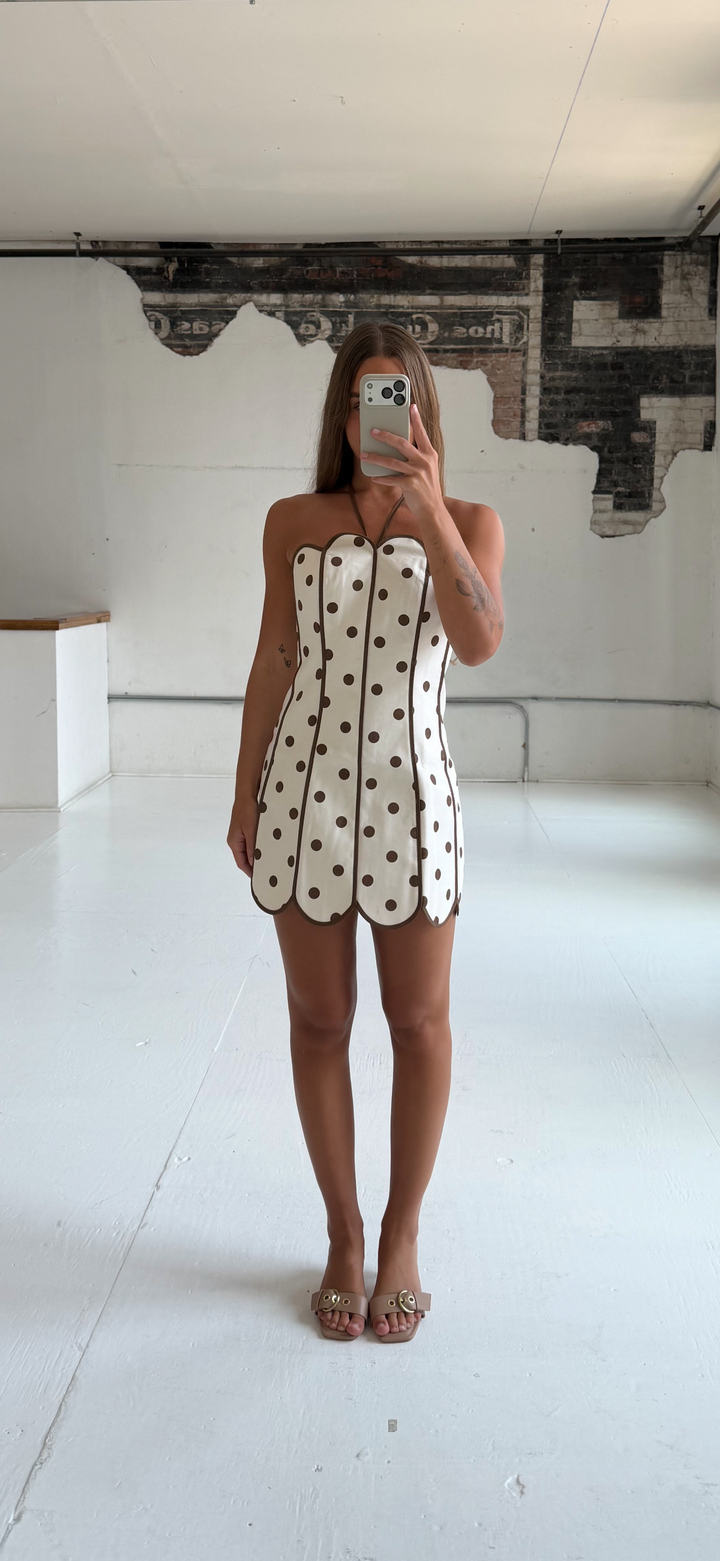 Woman wearing cream and brown polka dot mini dress with scallop neckline and hem.