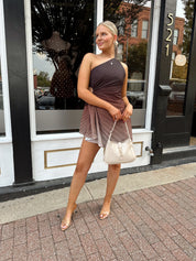 Woman wearing one shoulder brown ombré pleated mini dress with adjustable side cutout. 