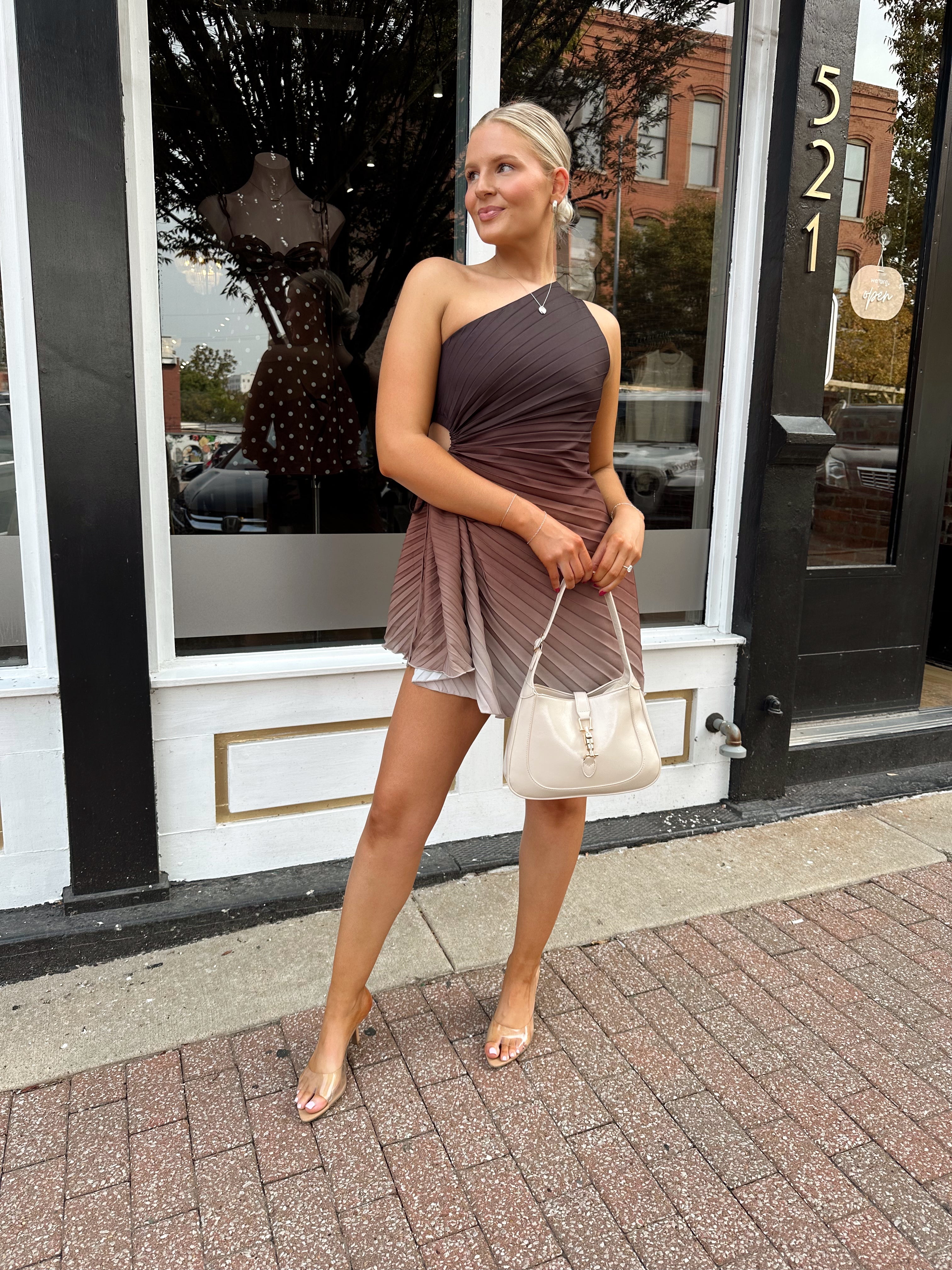 Woman wearing one shoulder brown ombré pleated mini dress with adjustable side cutout. 
