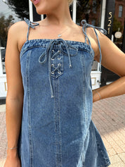 Woman wearing dark wash denim mini dress with lace up neckline detail.