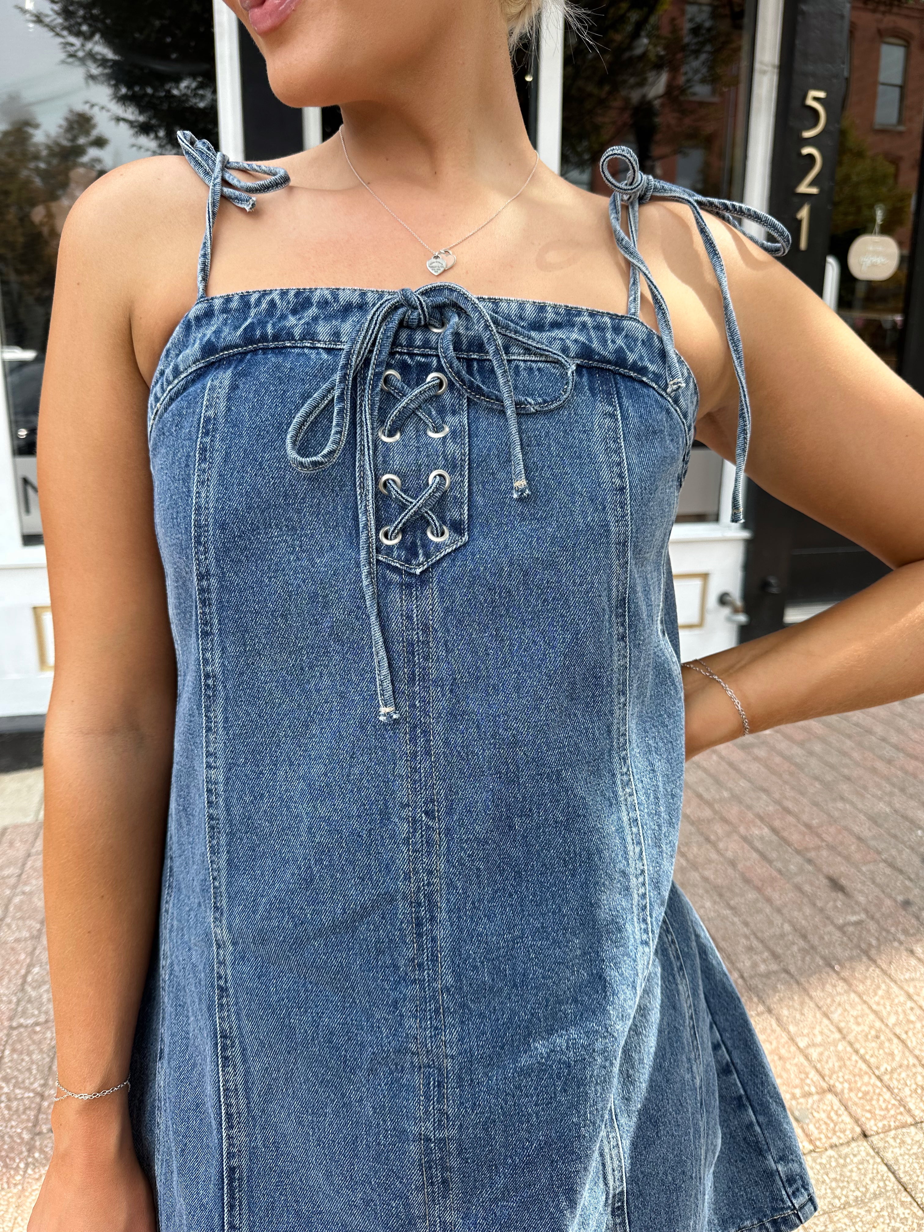 Woman wearing dark wash denim mini dress with lace up neckline detail.