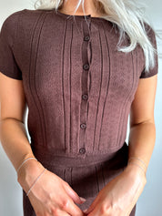 Woman with dark brown two piece knit set with short sleeve button down top, and midi length skirt with back slit.