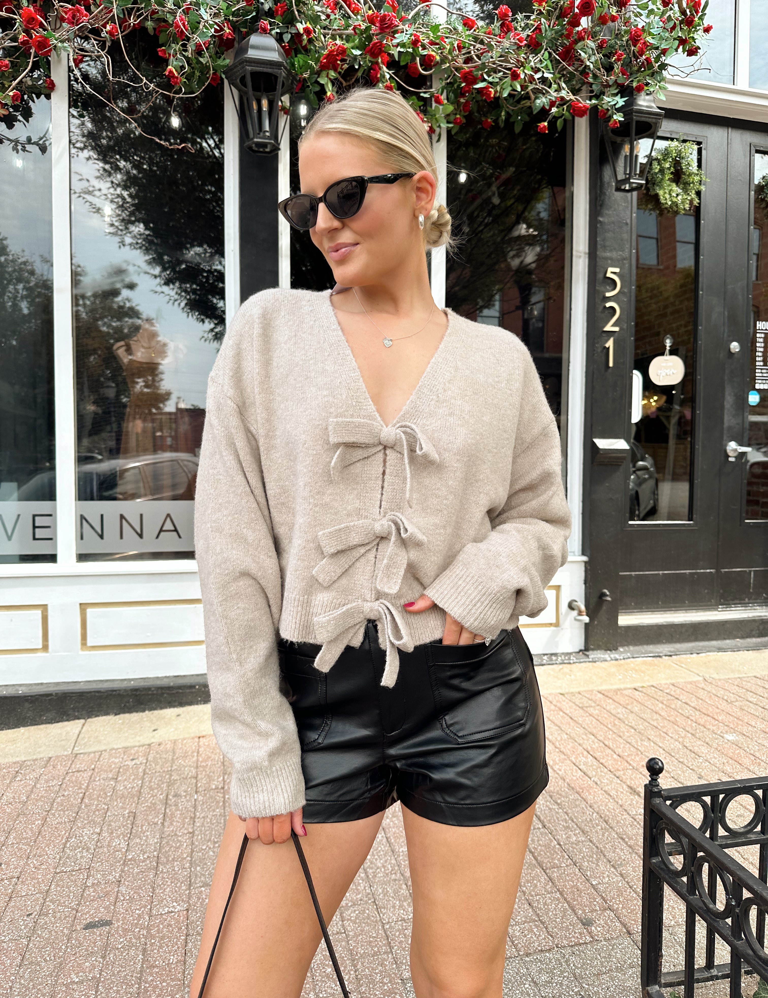 Woman wearing reversible taupe knit sweater with bow detailing and black leather short.