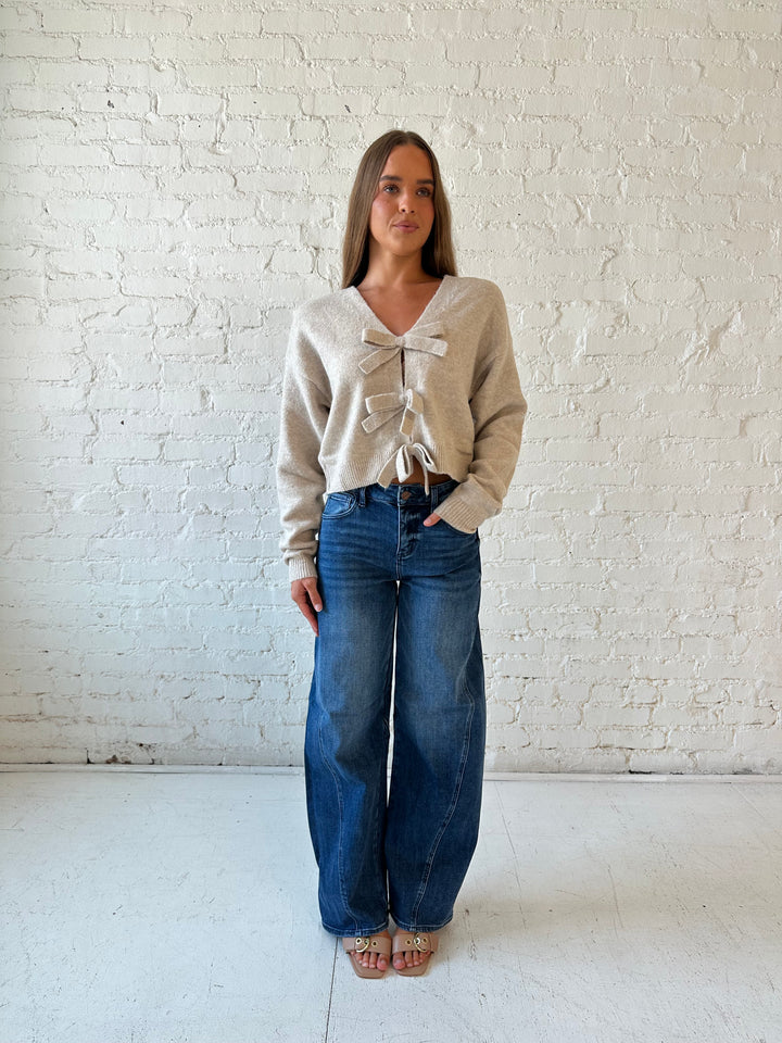 Woman wearing taupe knit reversible bow sweater with dark wash barrel jean.