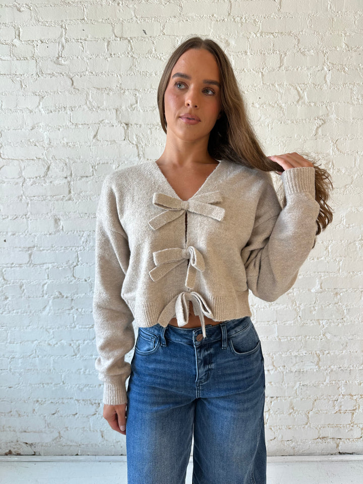Woman wearing taupe knit reversible bow sweater with dark wash barrel jean.