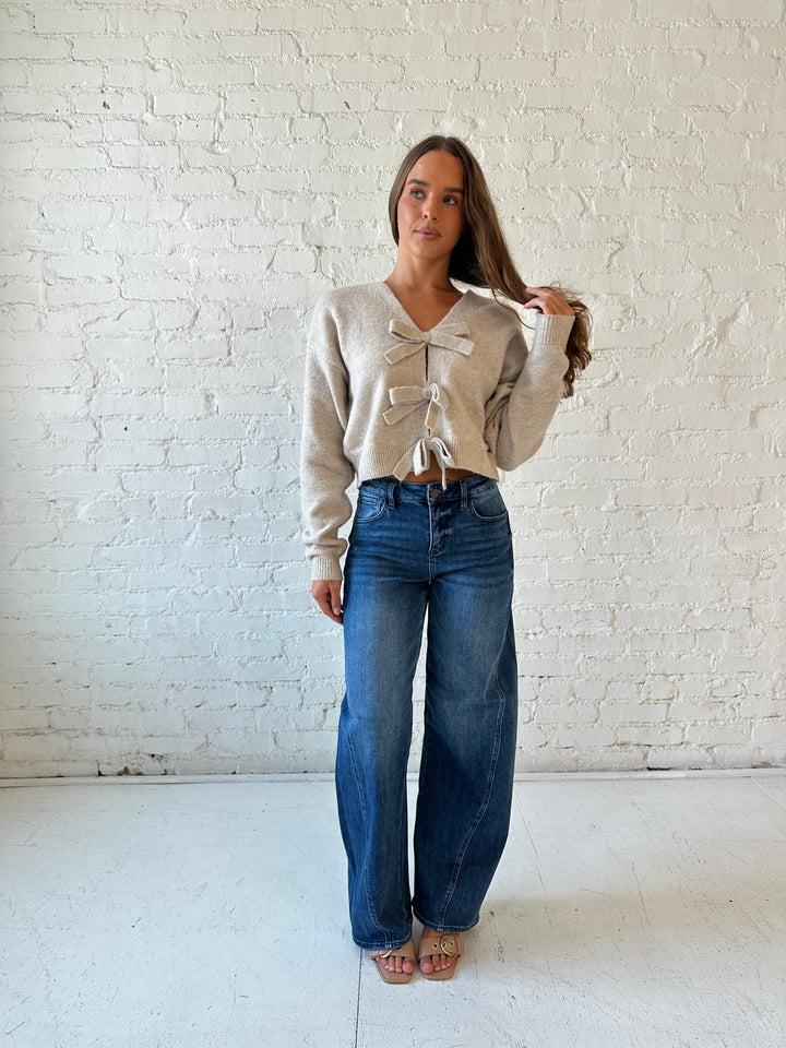 Woman wearing taupe knit reversible bow sweater with dark wash barrel jean.