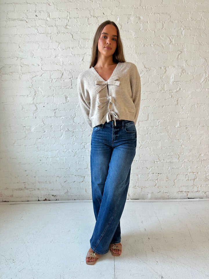 Woman wearing taupe knit reversible bow sweater with dark wash barrel jean.