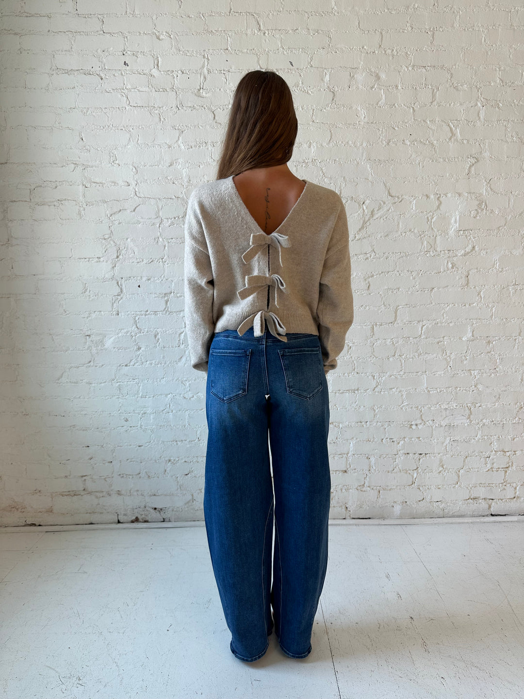 Woman wearing taupe knit reversible bow sweater with dark wash barrel jean.