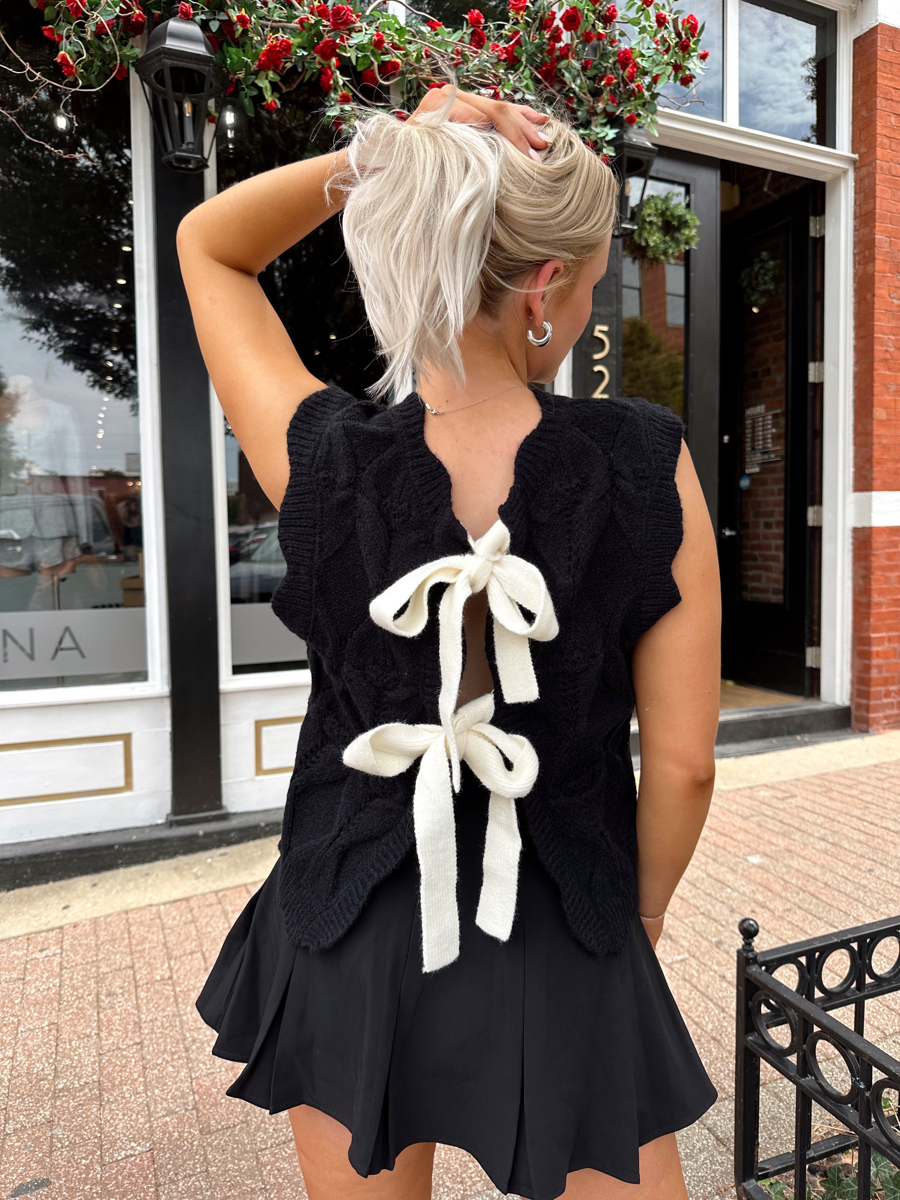 Woman wearing black sleeveless cable knit sweater with cream bow back.  