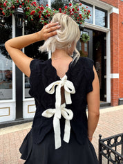 Woman wearing black sleeveless cable knit sweater with cream bow back.  