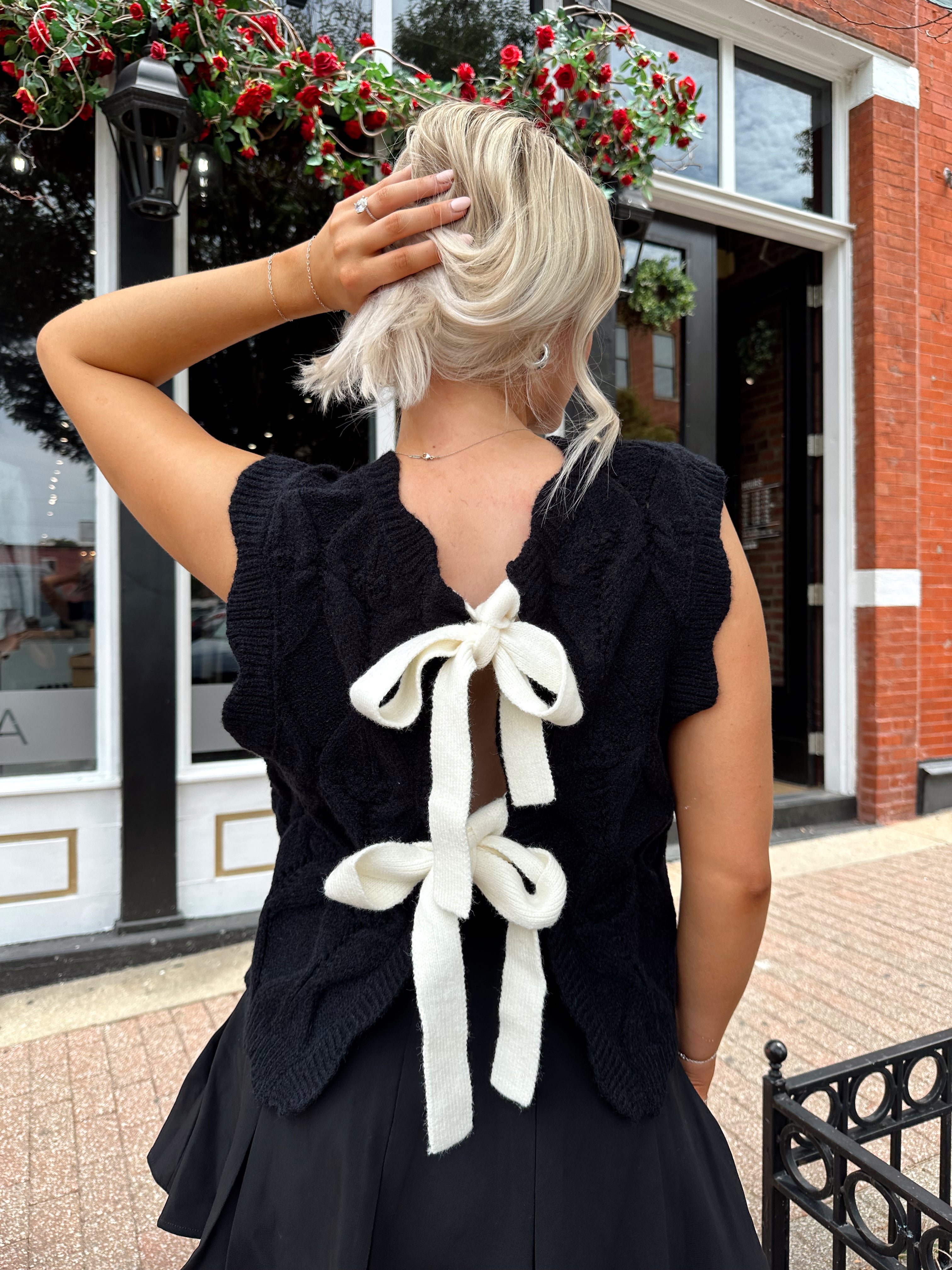 Woman wearing black sleeveless cable knit sweater with cream bow back.  