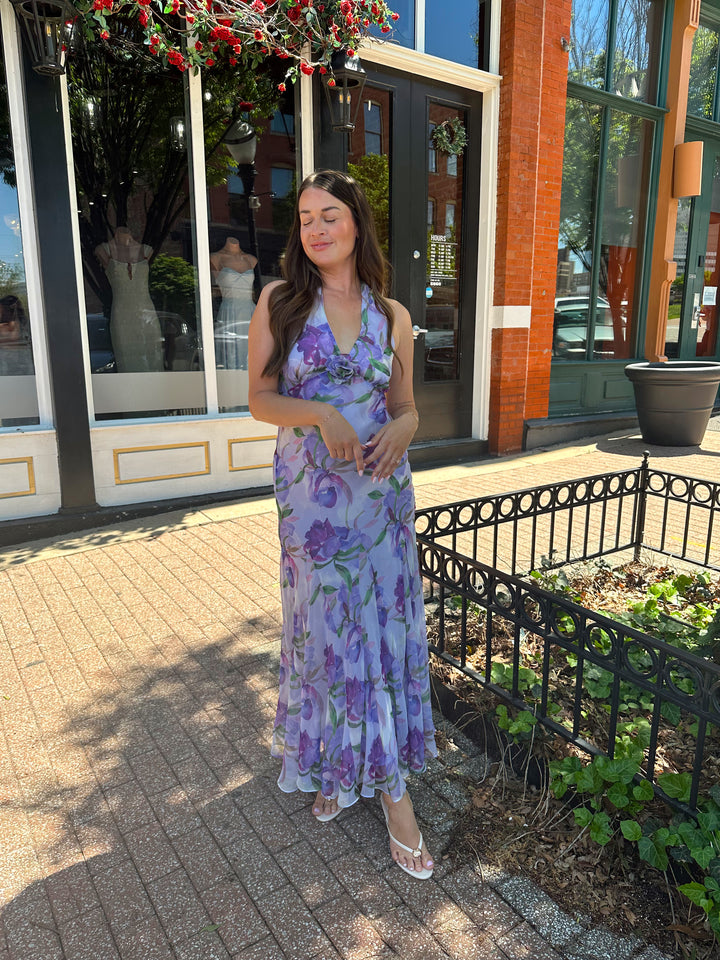 Woman wearing purple floral halter maxi dress with rosette detail. 