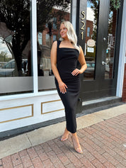 Woman wearing a black square neck fitted midi dress with clear heels outside a storefront.
