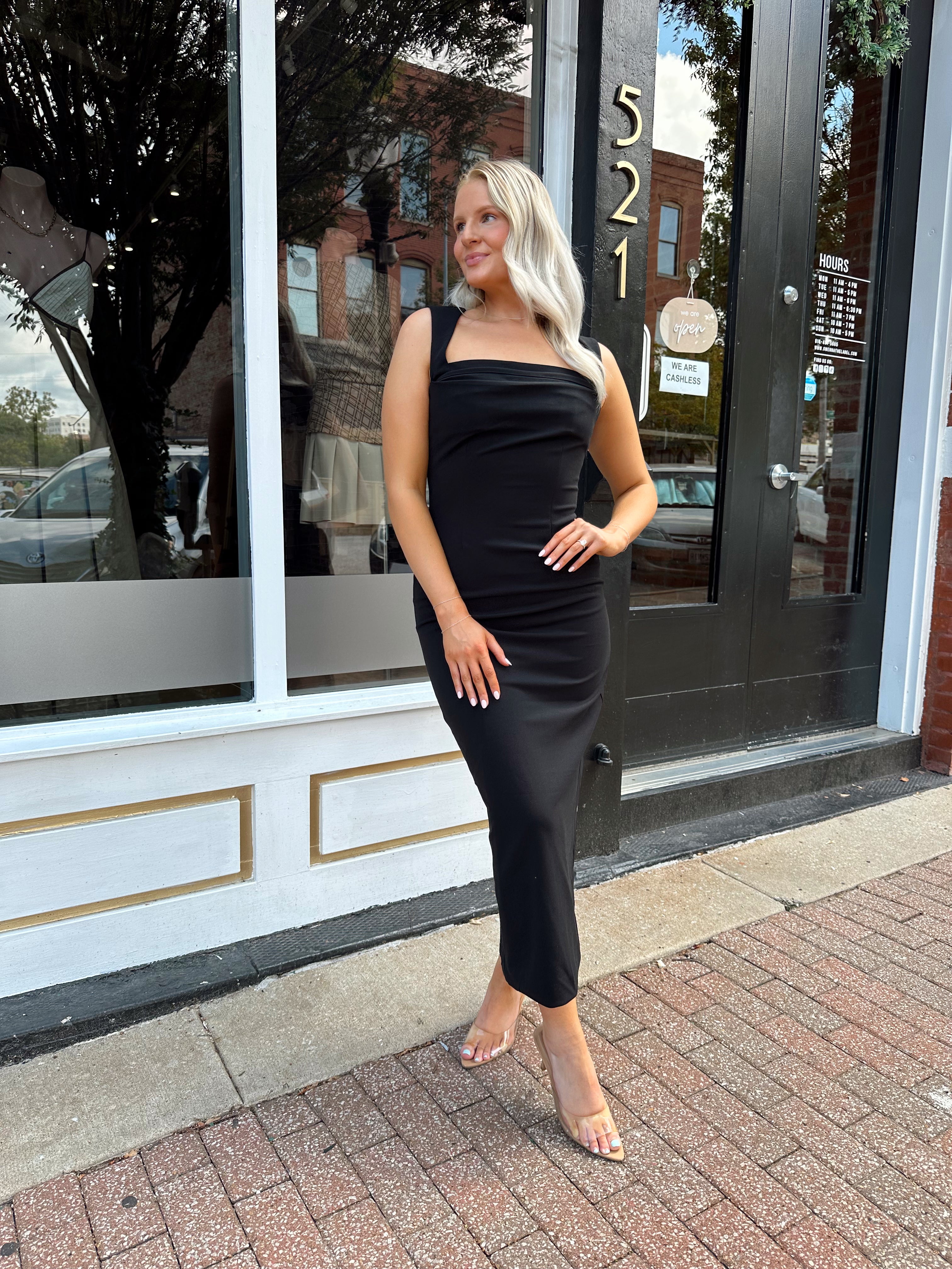 Woman wearing a black square neck fitted midi dress with clear heels outside a storefront.