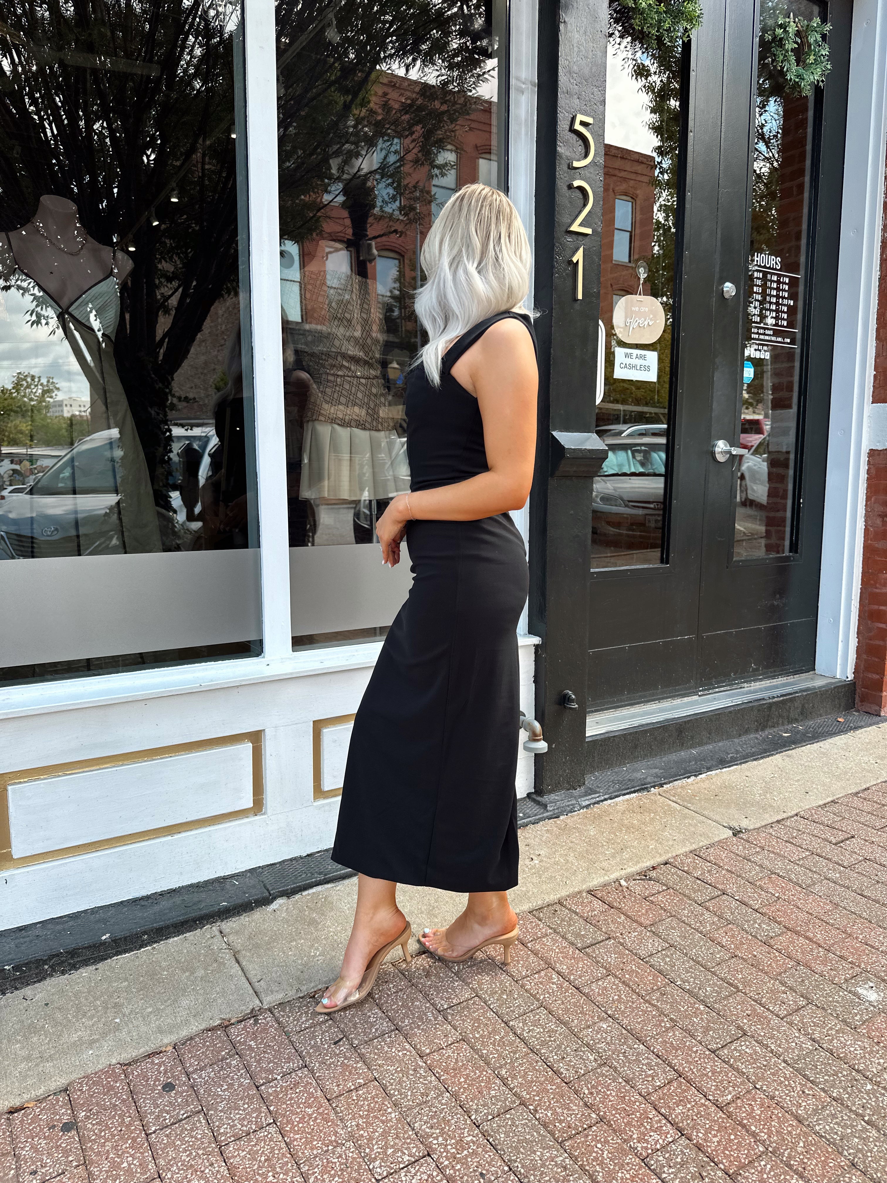 Woman wearing a black square neck fitted midi dress with clear heels outside a storefront.