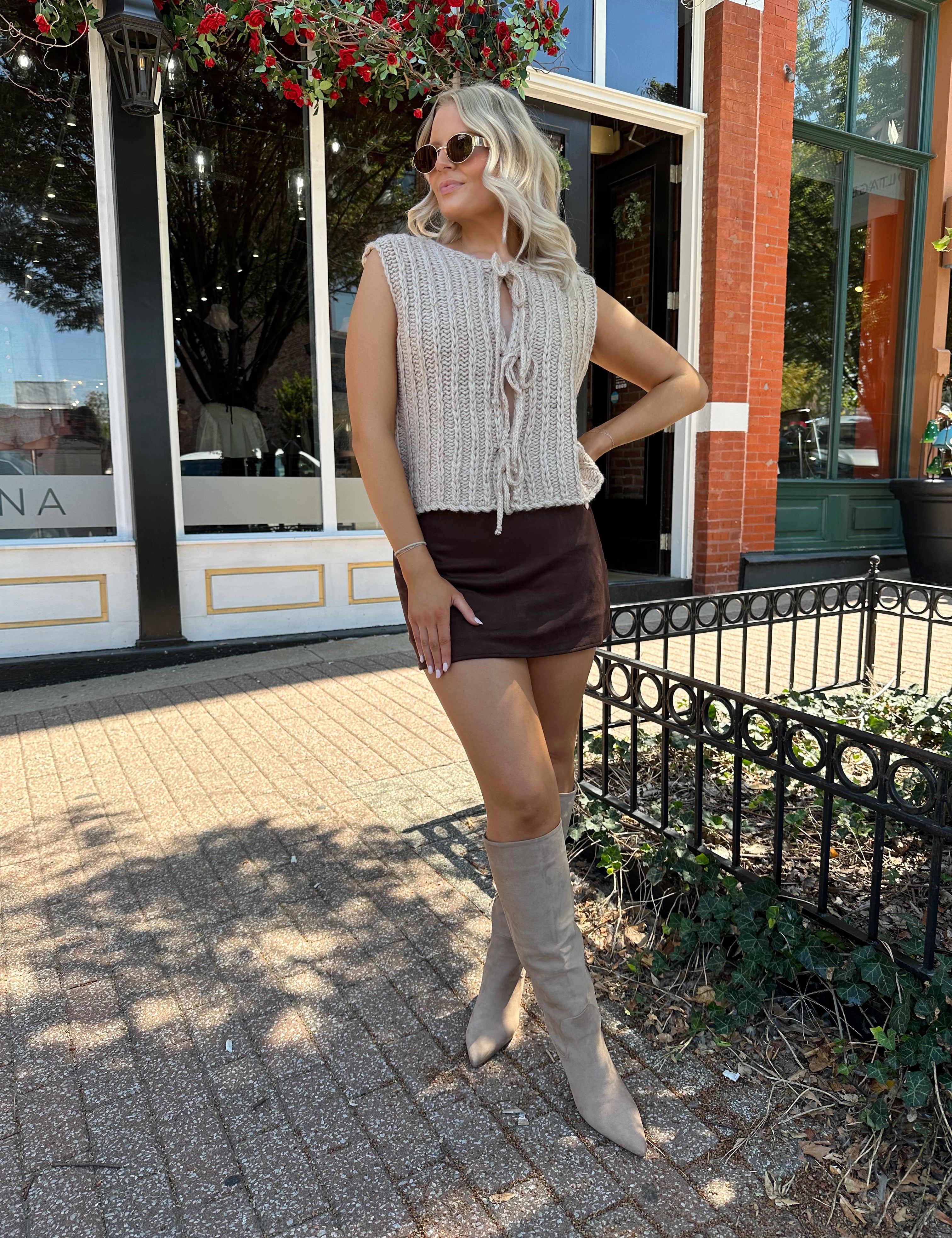 Woman wearing oatmeal colored chunky sleeveless sweater with front tie closure. 