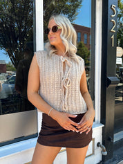 Woman wearing oatmeal colored chunky sleeveless sweater with front tie closure. 