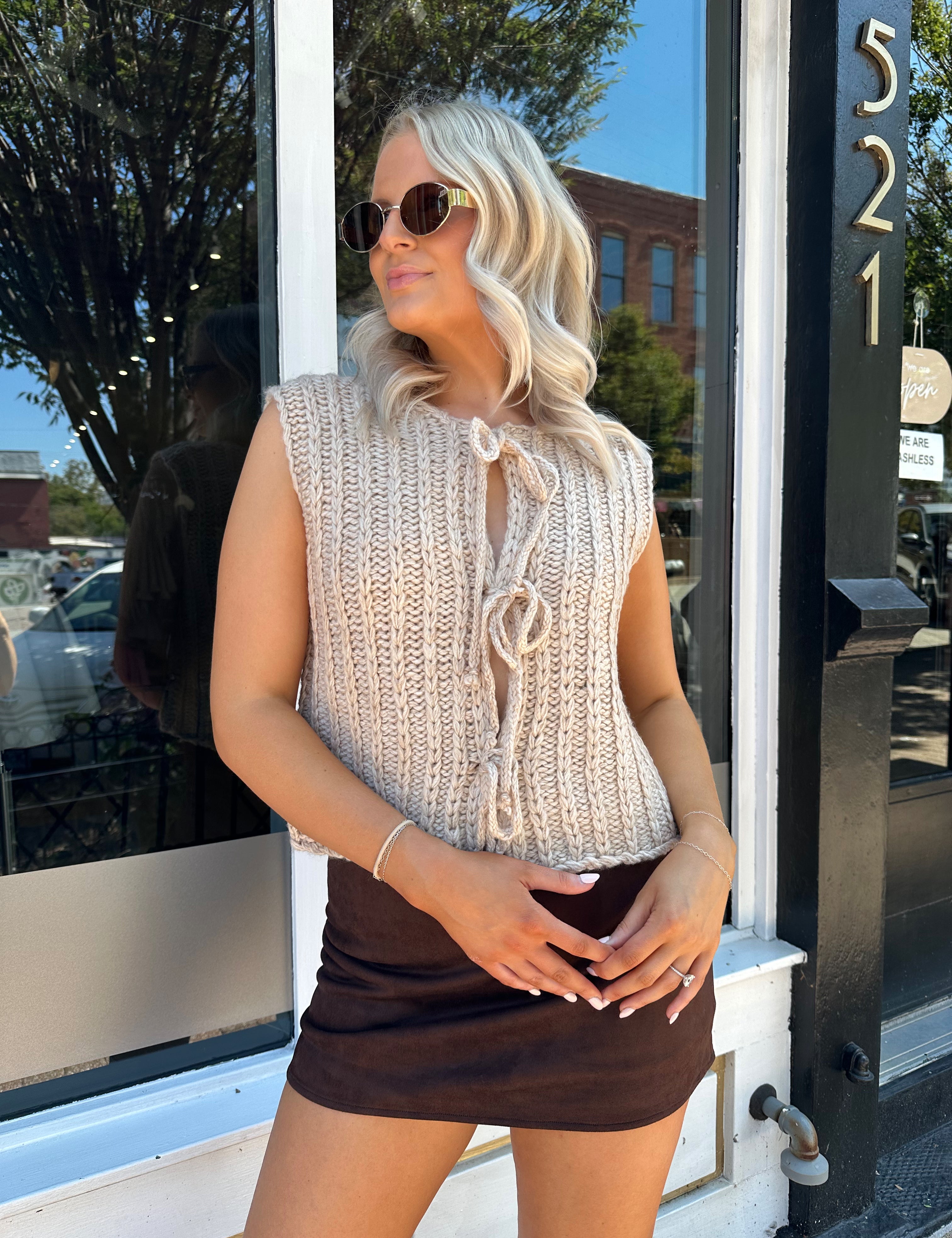 Woman wearing oatmeal colored chunky sleeveless sweater with front tie closure. 
