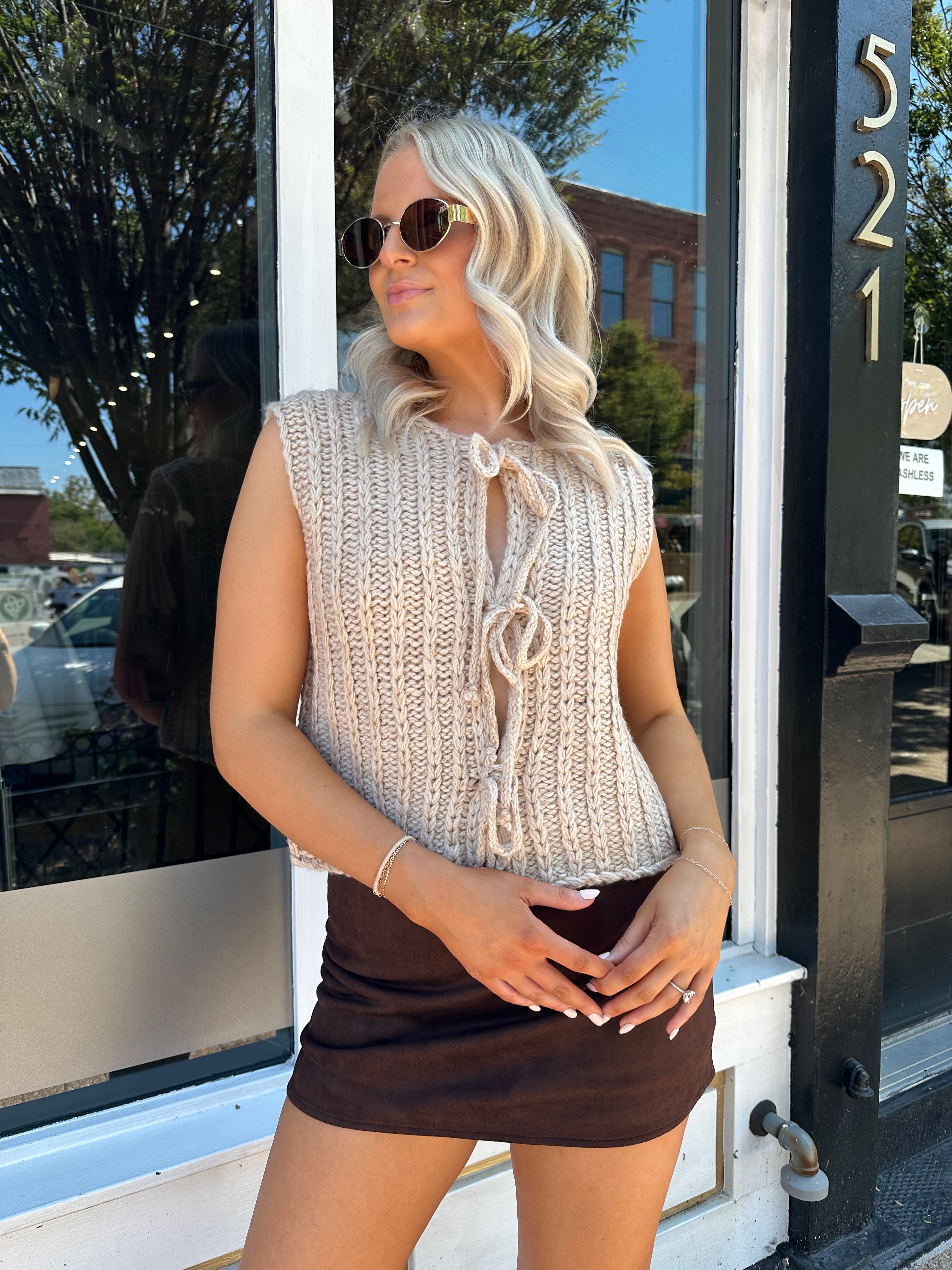 Woman wearing oatmeal colored chunky sleeveless sweater with front tie closure. 