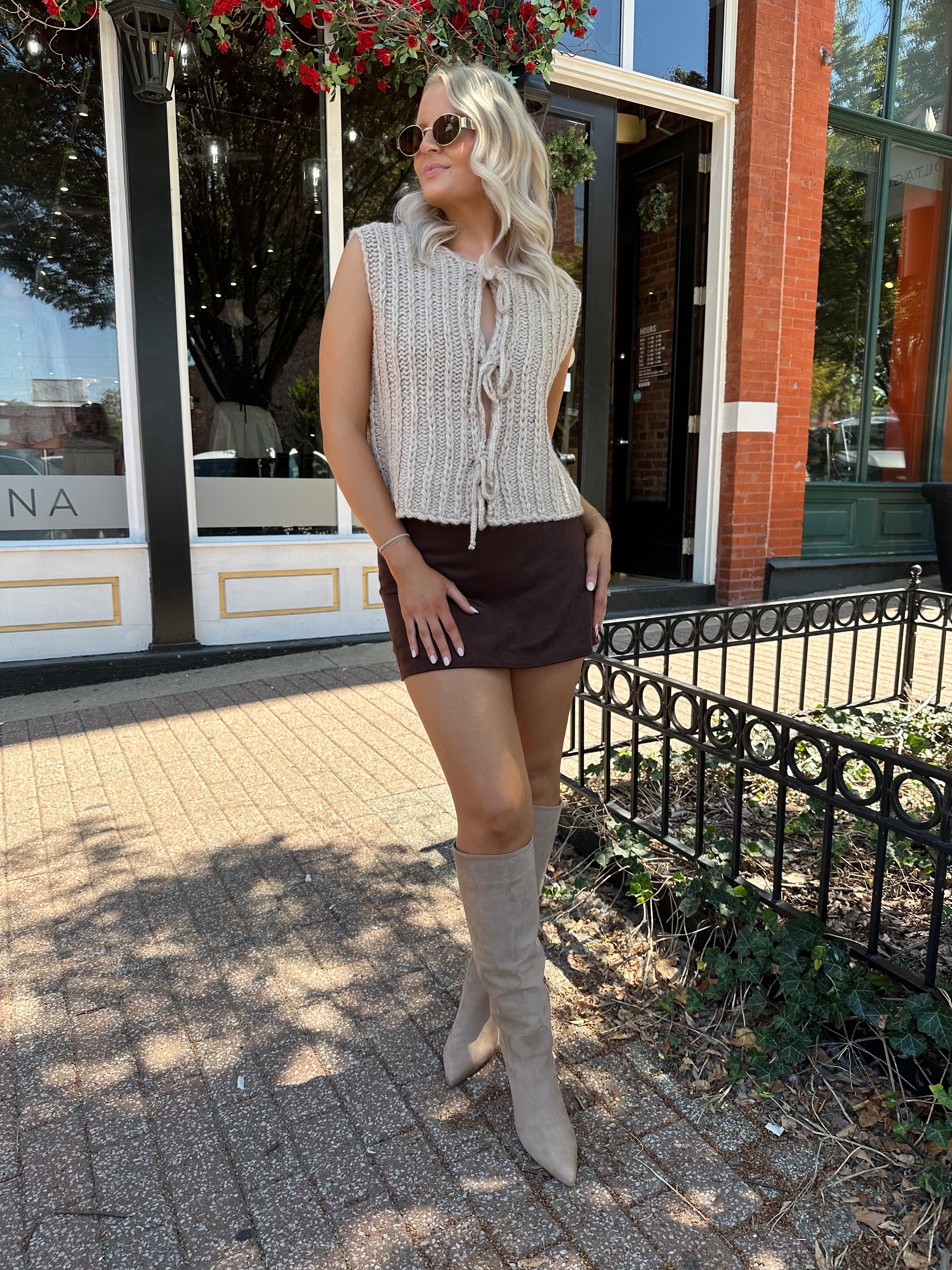 Woman wearing oatmeal colored chunky sleeveless sweater with front tie closure. 