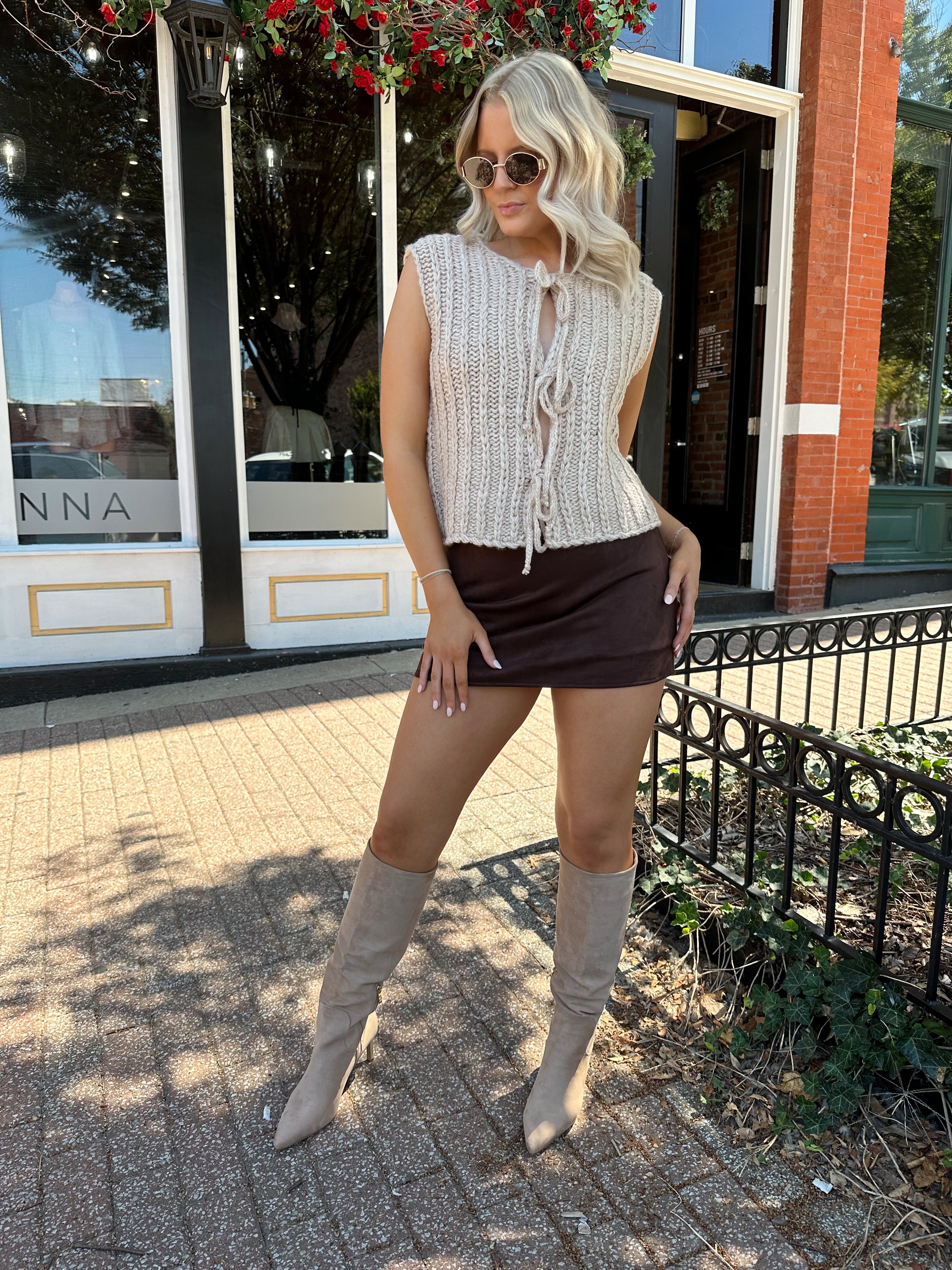 Woman wearing oatmeal colored chunky sleeveless sweater with front tie closure. 