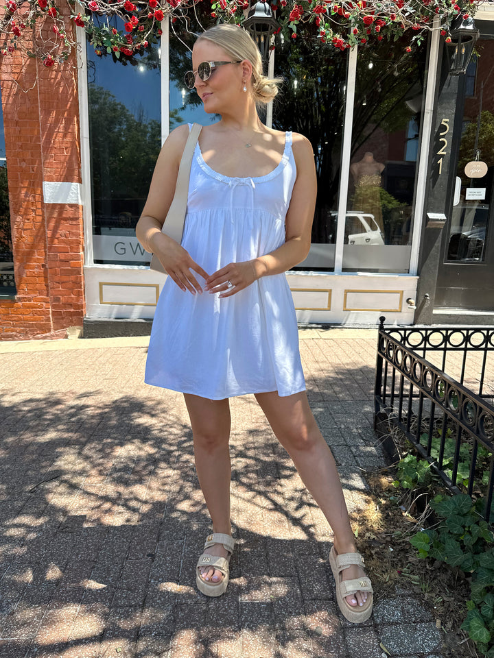 Woman wearing white mini dress with scoop neckline, ruffle trim, and functional pockets. 