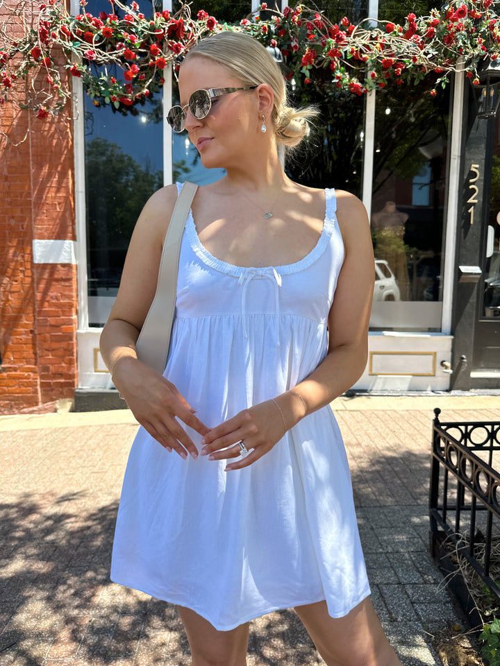 Woman wearing white mini dress with scoop neckline, ruffle trim, and functional pockets. 