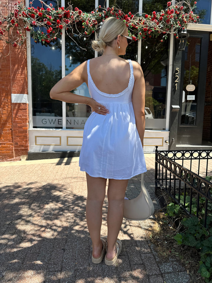 Woman wearing white mini dress with scoop neckline, ruffle trim, and functional pockets. 