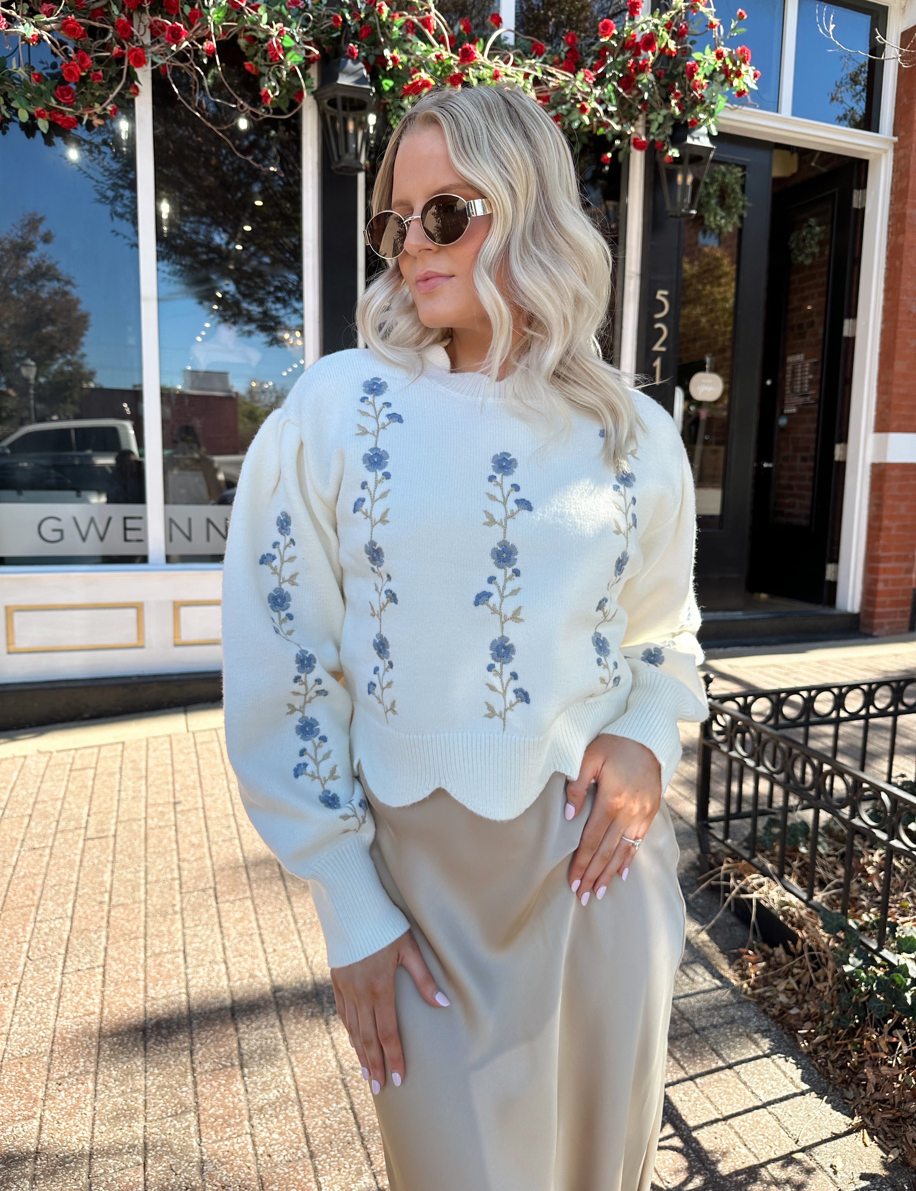 Woman wearing cream sweater with blue embroidered floral pattern. 