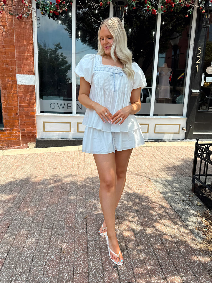Woman wearing white two piece set with a puff sleeve babydoll top and flowy shorts. 