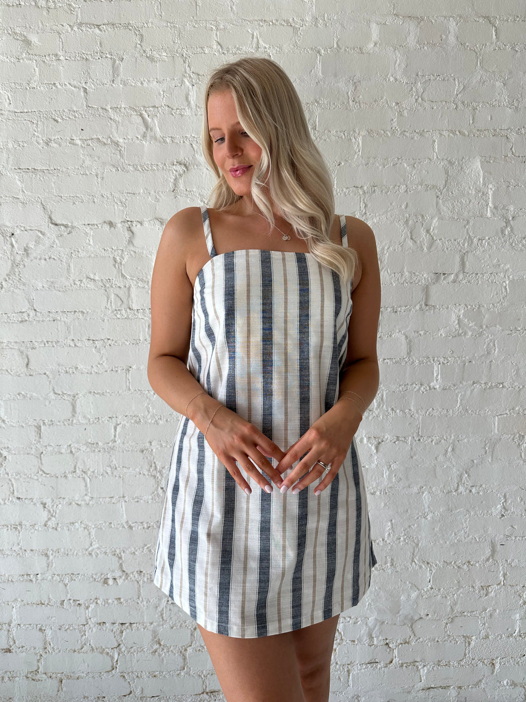Woman wearing neutral striped linen mini dress with straight neckline and adjustable straps. 