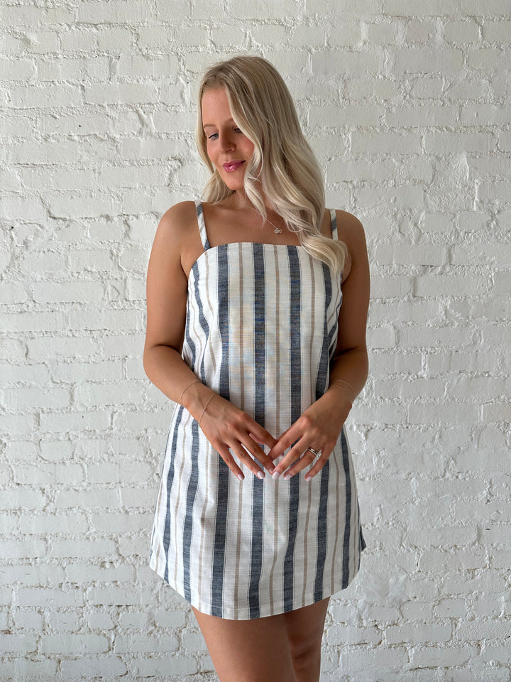 Woman wearing neutral striped linen mini dress with straight neckline and adjustable straps. 