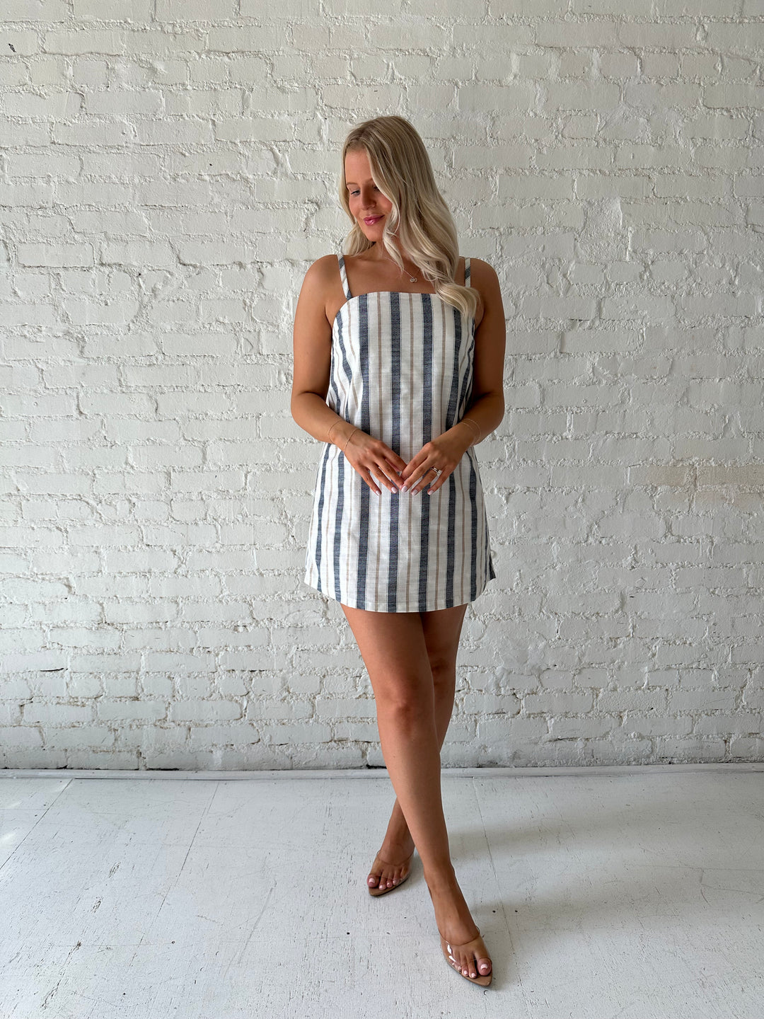 Woman wearing neutral striped linen mini dress with straight neckline and adjustable straps. 