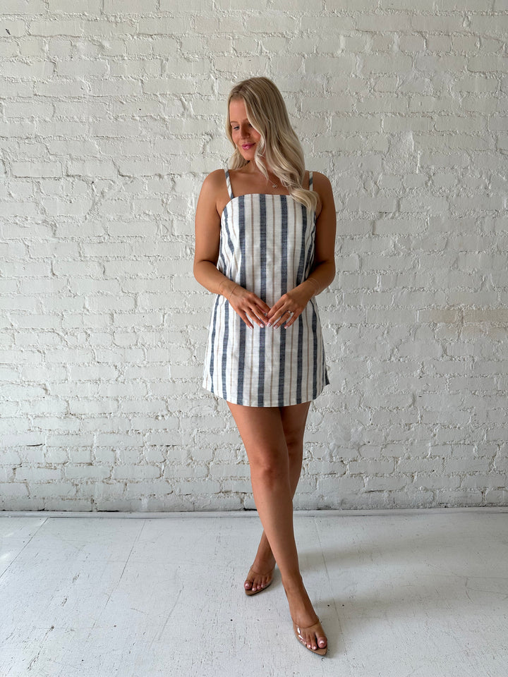 Woman wearing neutral striped linen mini dress with straight neckline and adjustable straps. 