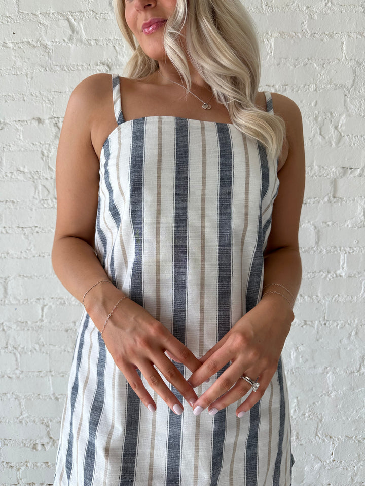 Woman wearing neutral striped linen mini dress with straight neckline and adjustable straps. 